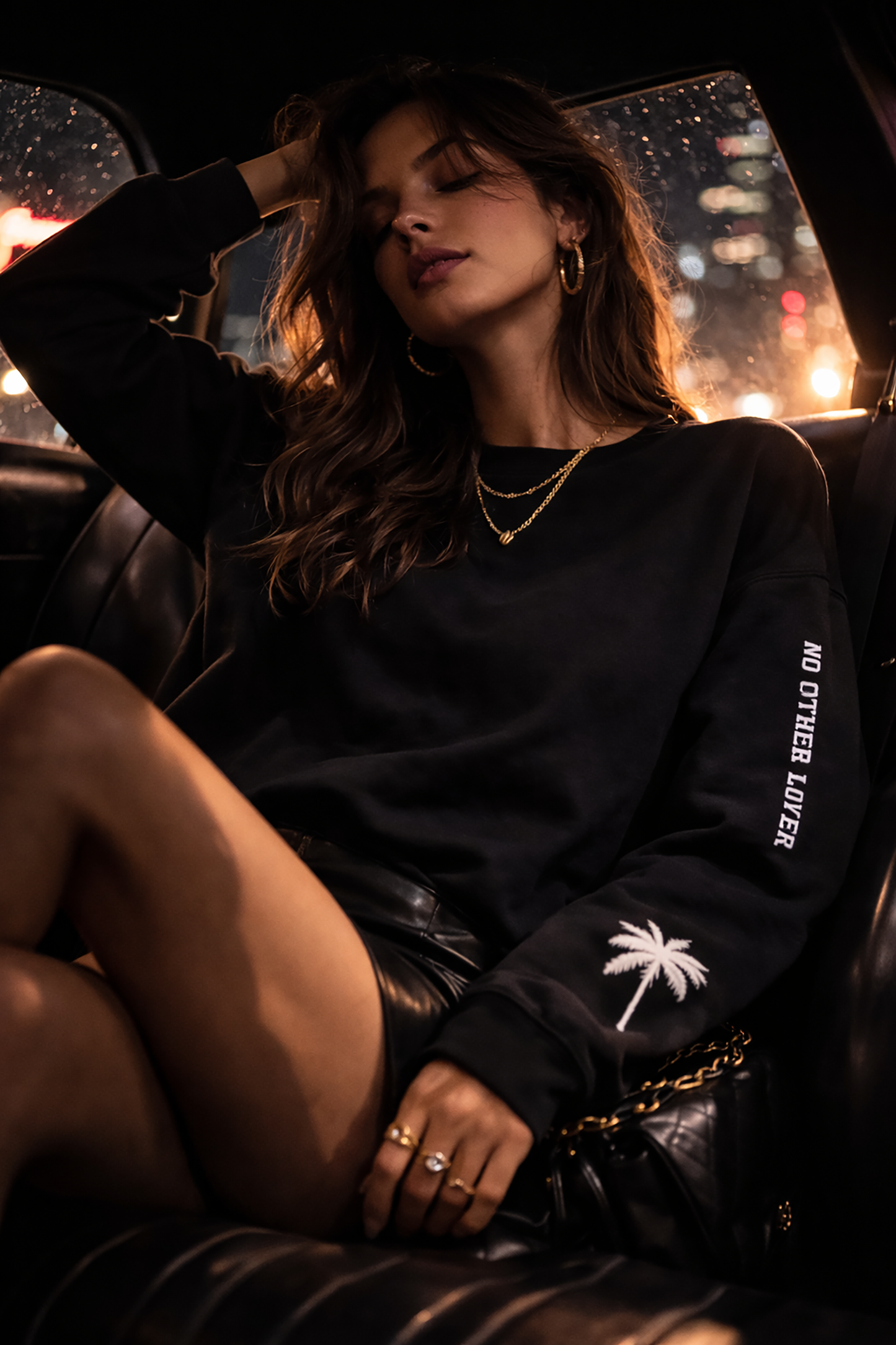Woman sitting inside a car at night wearing a No Other Lover logo and palm tree design on the sleeves, with city lights in the background