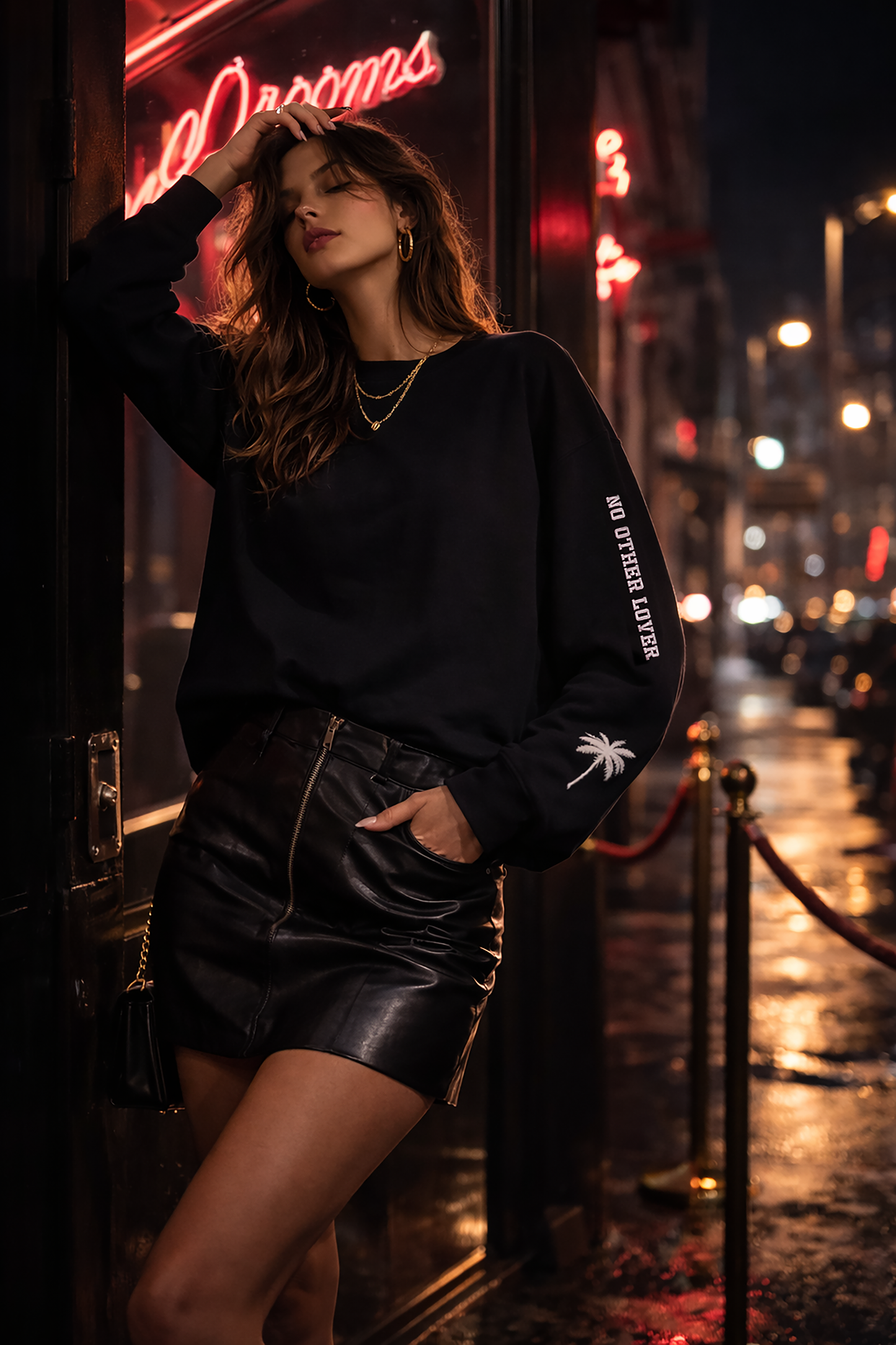 Woman in a black No Other Lover logo and palm tree sleeves sweatshirt, and leather skirt standing on a city street at night.