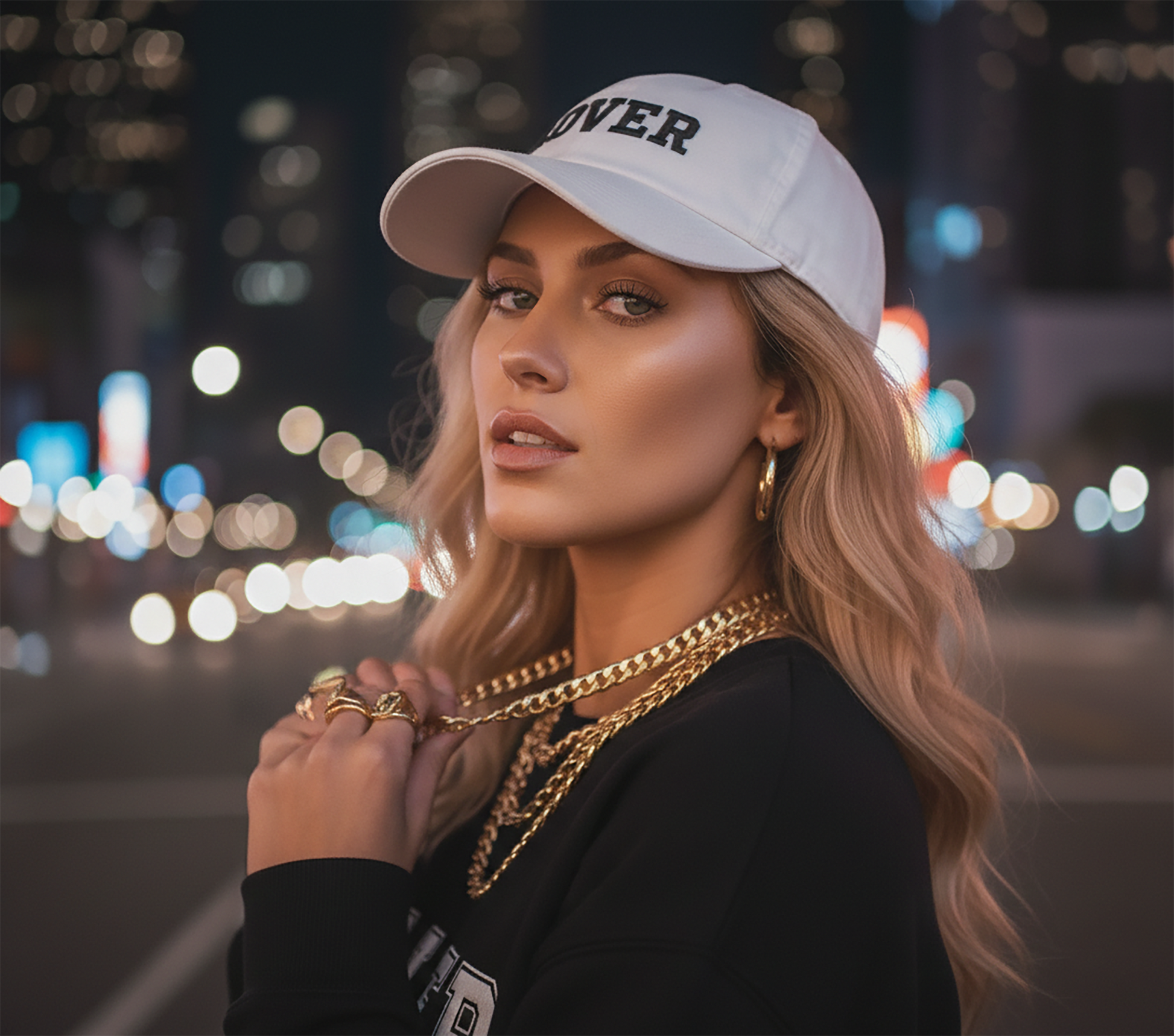 Woman wearing a No Other Lover LOVER White Cotton Baseball Cap, gold chain necklace, and black LOVER sweatshirt against a blurred city lights background.