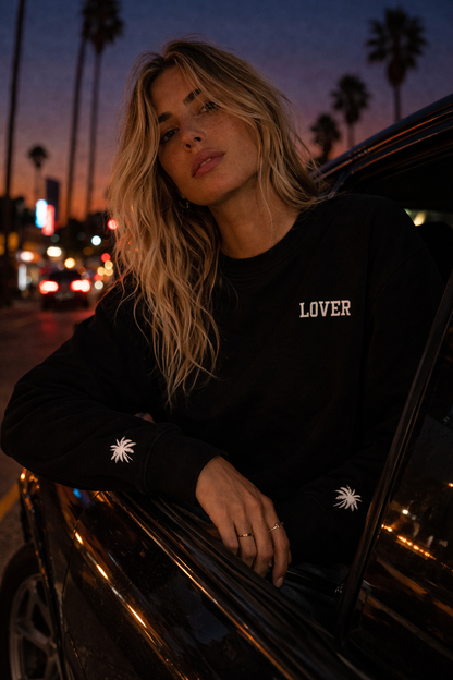 Woman wearing a black No Other Lover LOVER Palms Sweatshirt with 'LOVER' text, leaning out of a car at night with palm trees in the background.