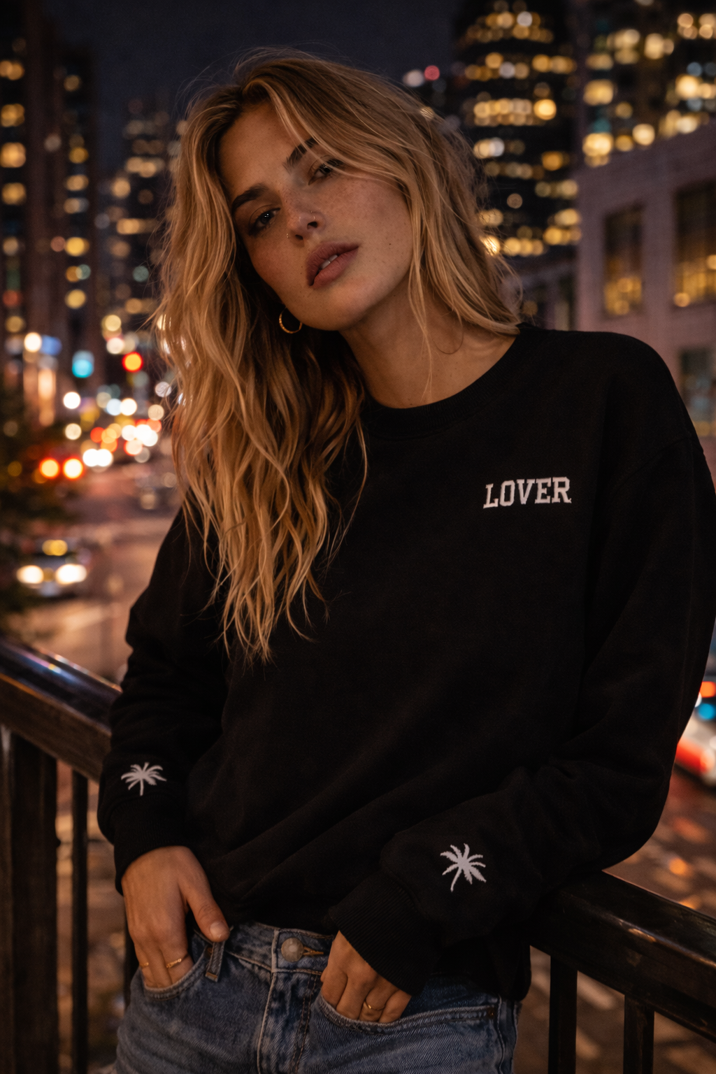 Woman wearing a black No Other Lover LOVER Palms Sweatshirt with 'LOVER' text, on a balcony against a cityscape at night.
