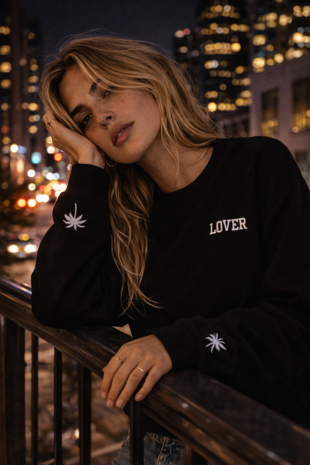 Woman wearing a black No Other Lover LOVER Palms Sweatshirt with 'LOVER' text and palm tree designs, standing against a cityscape at night.