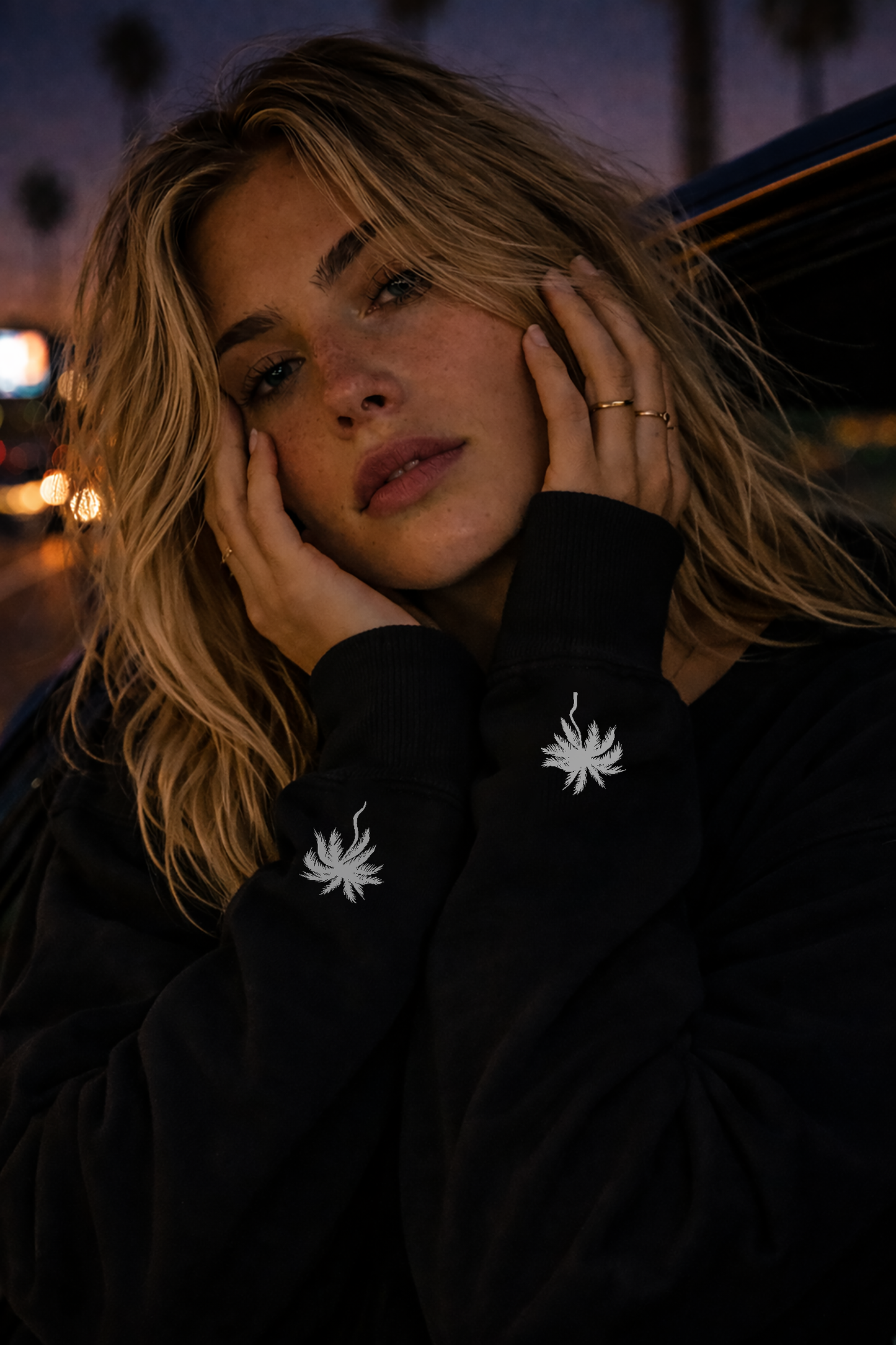 Woman wearing a black No Other Lover LOVER Palms Sweatshirt with white palm tree designs, leaning out of a car window at night.