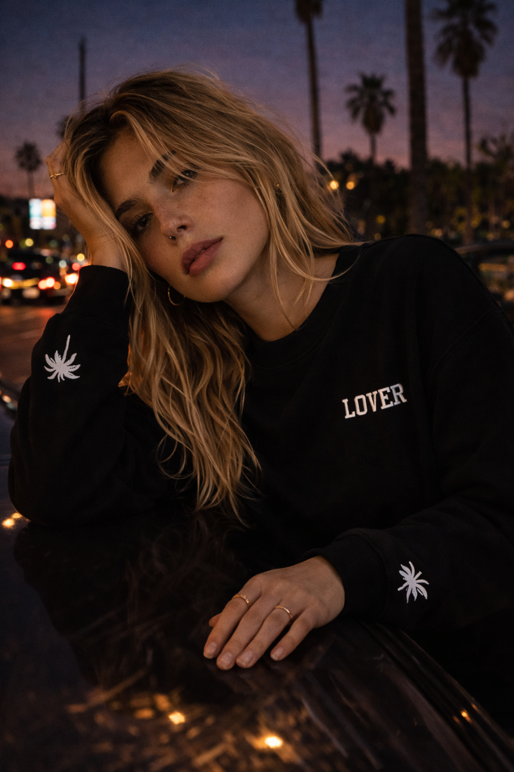 Woman wearing a black No Other Lover LOVER Palms Sweatshirt with palm trees and city lights in the background