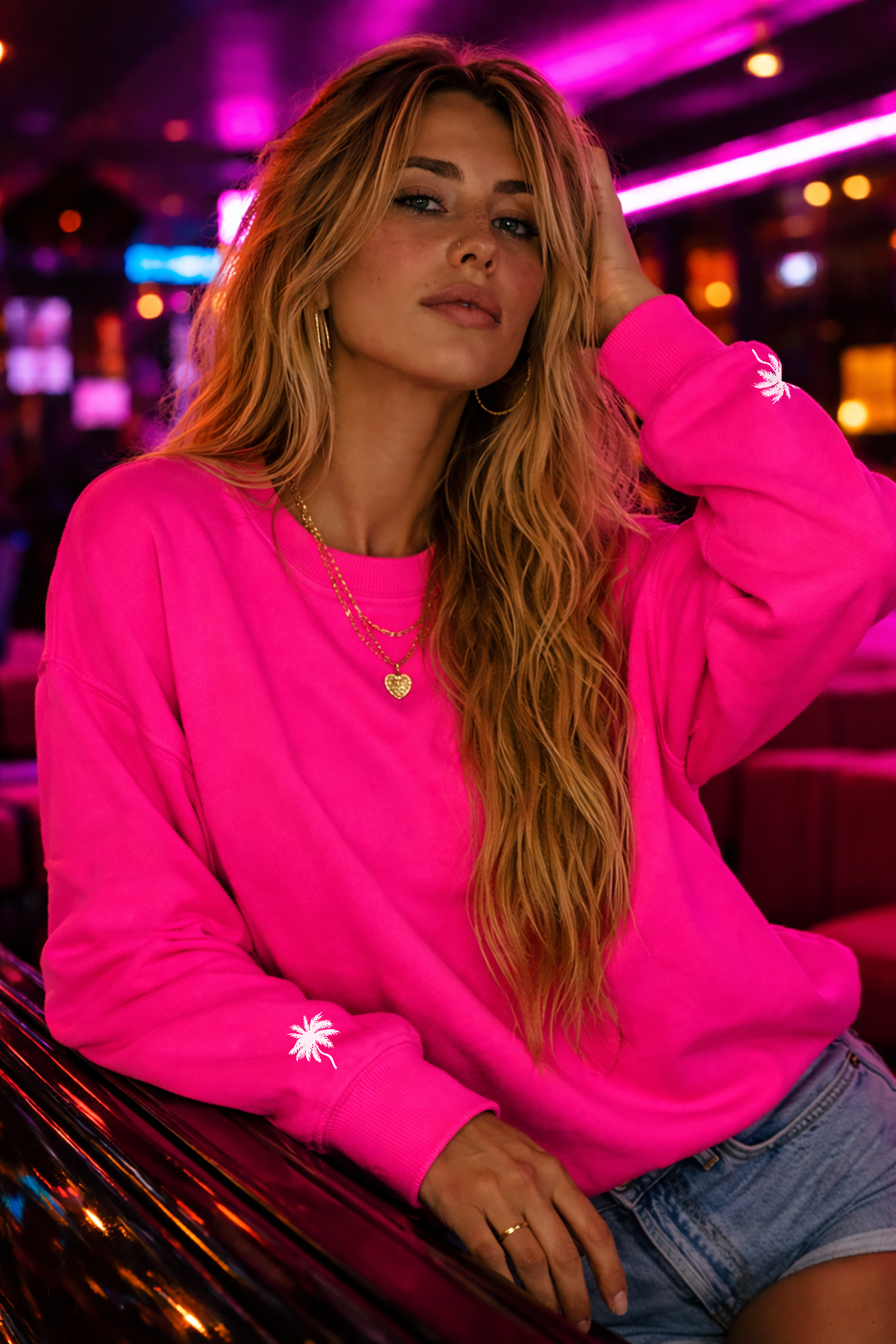Woman wearing a hot pink No Other Lover LOVER Palms Sweatshirt with palm tree designs on the cuffs in a neon-lit indoor setting.