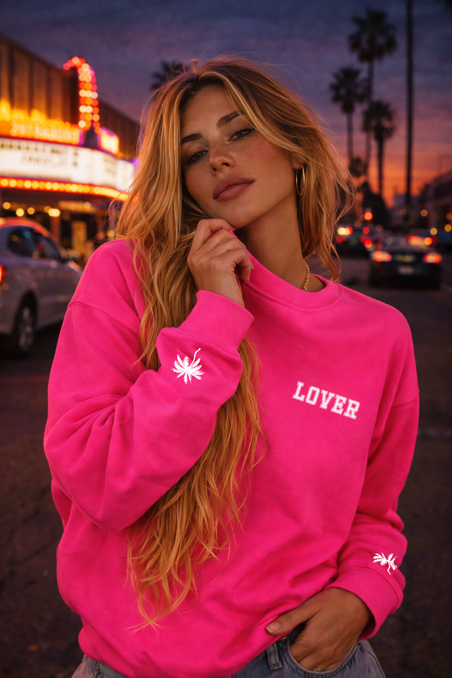 Woman wearing a hot pink No Other Lover LOVER Palms Sweatshirt with 'LOVER' text at sunset.