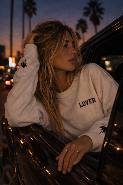 Blonde woman wearing a white No Other Lover LOVER Palms retro 80s sweatshirt with palm sleeve detail leaning out of a car  window at night with palm trees and city lights in the background.