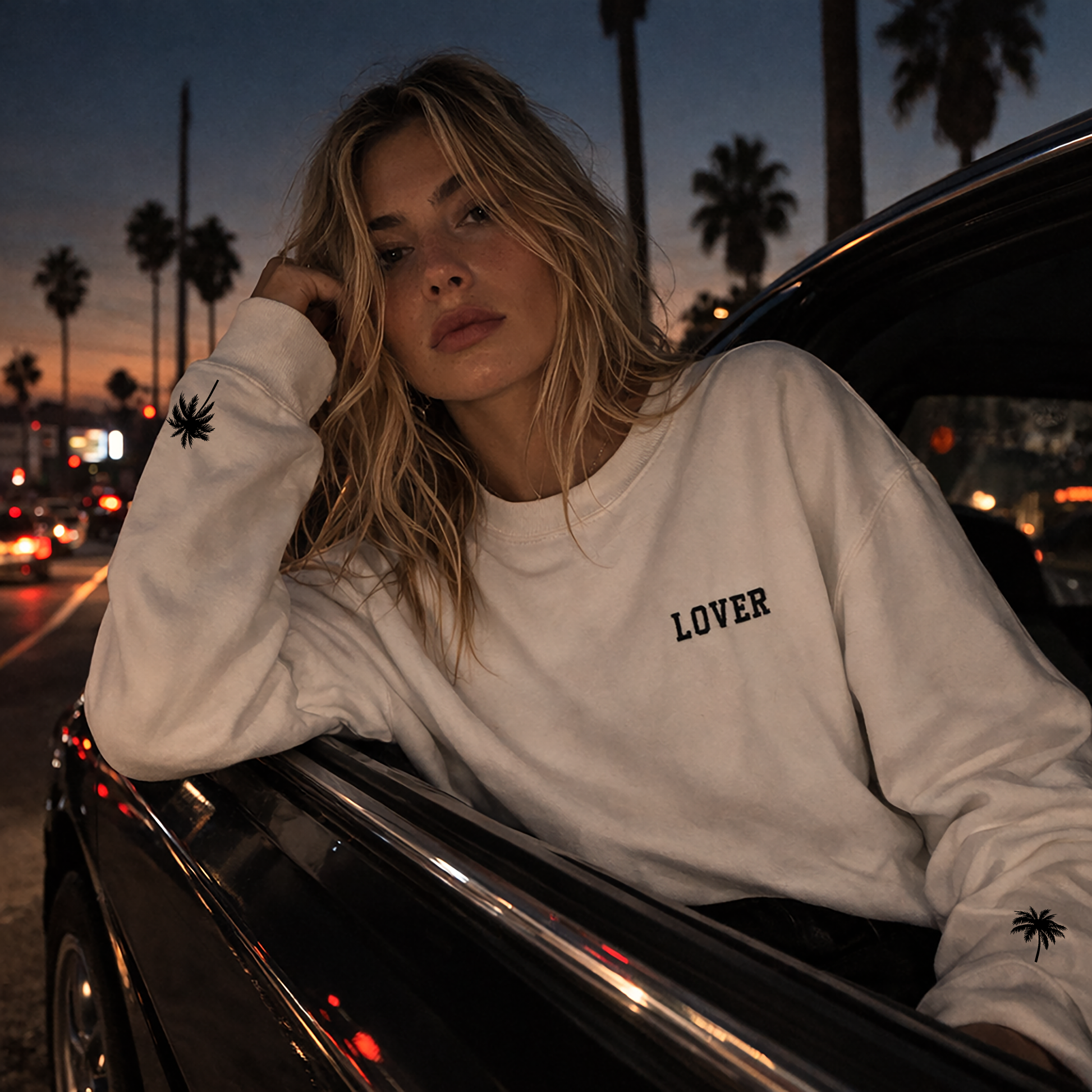 Woman wearing a white No Other Lover LOVER Palms retro 80s sweatshirt with palm sleeve detail, leaning against a car at dusk with palm trees in the background.