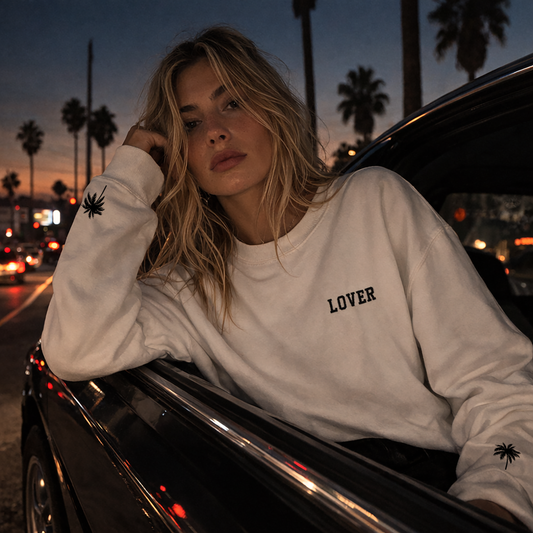 Woman wearing a white No Other Lover LOVER Palms retro 80s sweatshirt with palm sleeve detail, leaning against a car at dusk with palm trees in the background.