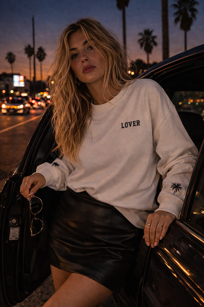 Woman in a No Other Lover LOVER Palms retro 80s sweatshirt with palm sleeve detail leaning against a car at dusk with palm trees and city lights in the background.
