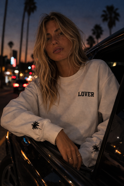 Woman wearing a white No Other Lover LOVER Palms retro 80s sweatshirt with palm sleeve detail, leaning against a car at night with palm trees and city streets in the background.